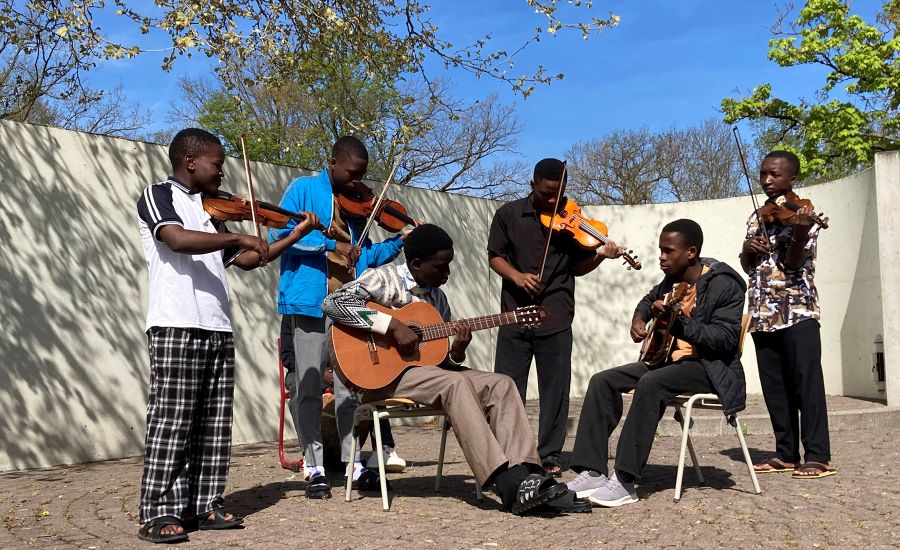Sechs Schüler aus Tansania spielen vor einer Mauer auf Geigen und Gitarren