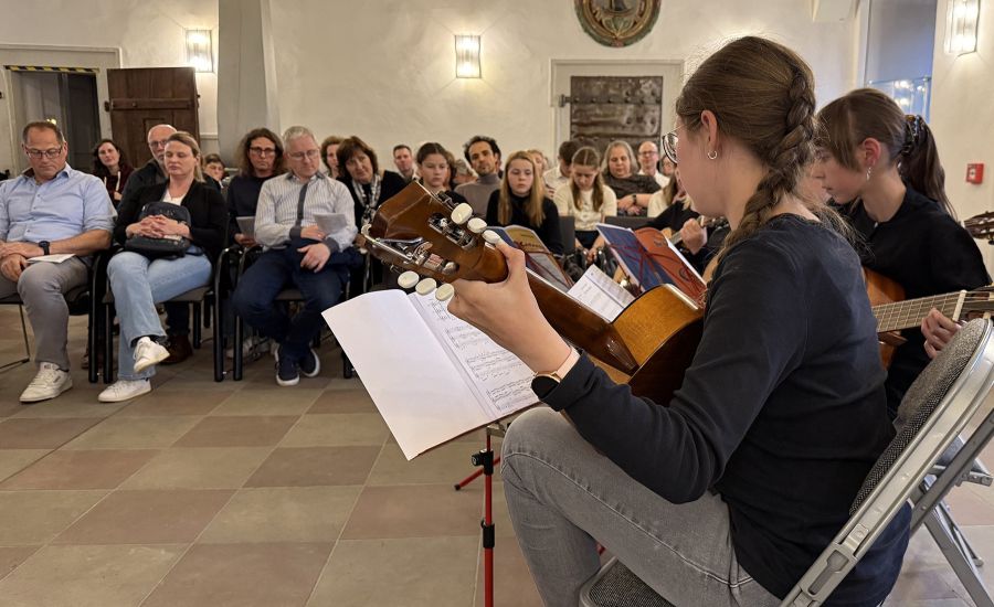 Zwei Mädchen mit Gitarre vor einem größeren Publikum auf Stühlen