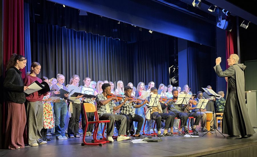 Sechs schwarze Schüler mit Geigen und Gitarren auf der Bühne, hinter ihn deutsche Schülerinnen und Schüler